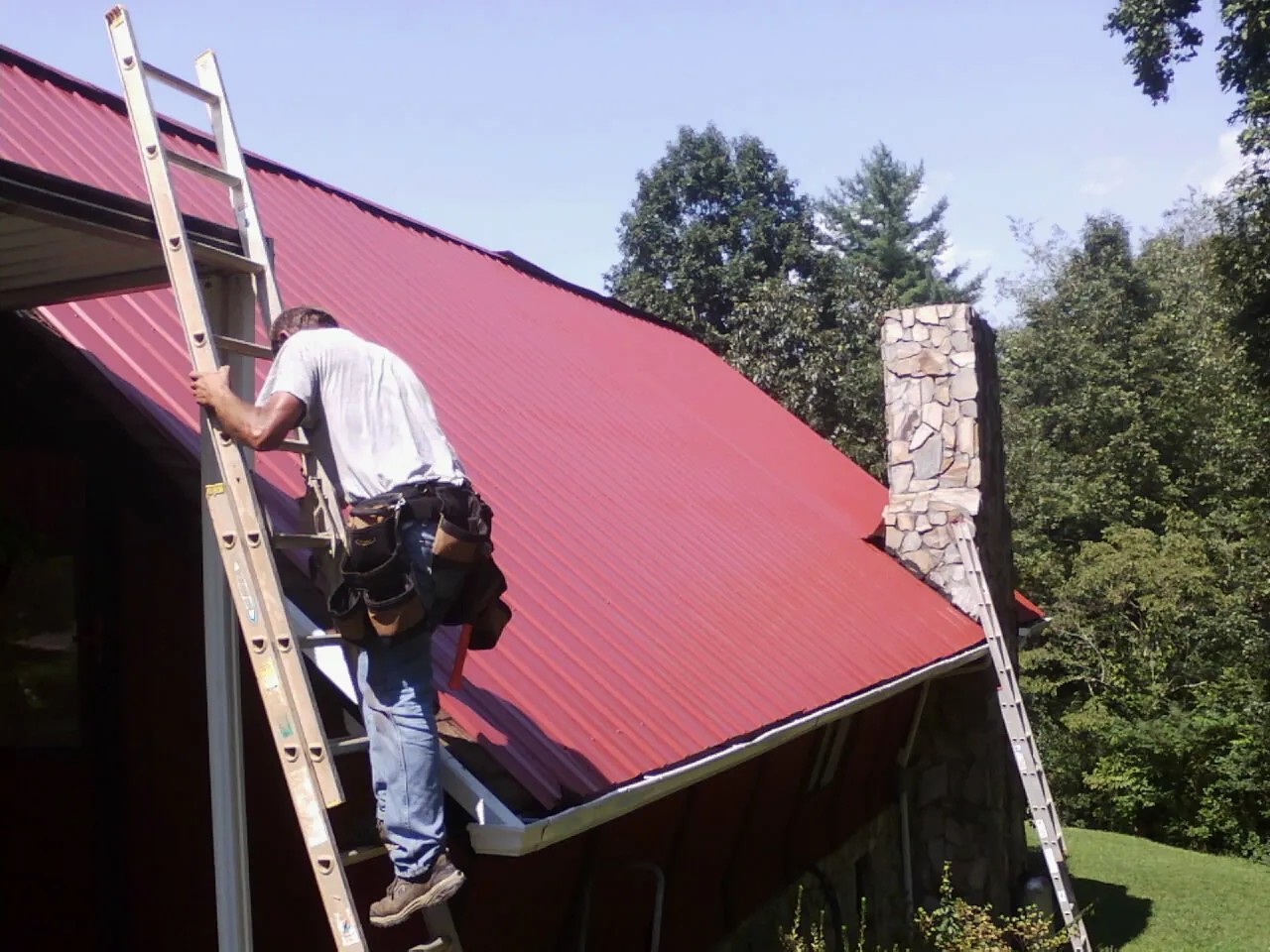 Roofer on a ladder beside a red metal roof and stone chimney — Soffit & Fascia Repair by Roofing Brothers in Seeley Lake